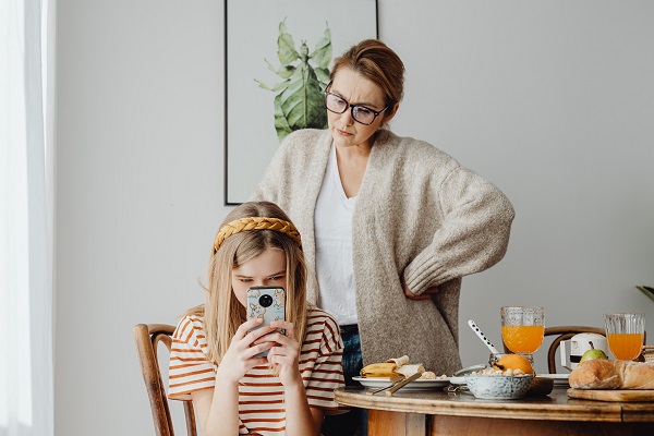 mama care sta in picioare si se uita cu atentie la fiica ei adolescenta, care se uita in telefon