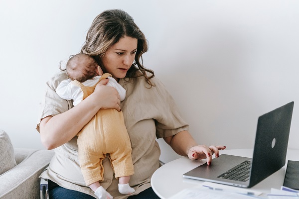 mama care isi tine bebelusul in brate in timp ce lucreaza pe laptop