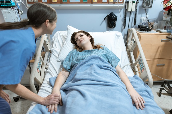 femeie tanara, asistenta medicala sau medic, care strange mana unei paciente aflate pe patul de spital