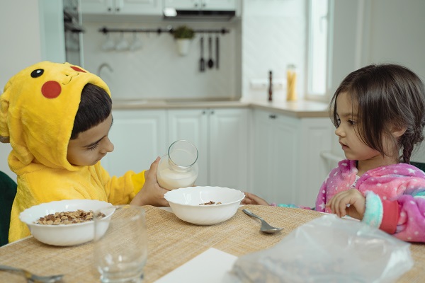 baietel imbracat in costum de Pikachu turnand lapte peste cerealele unei fetite cu care sta la masa