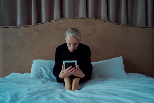 femeie in varsta imbracata in negru stand ghemuita pe pat si uitandu-se la o fotografie inramata