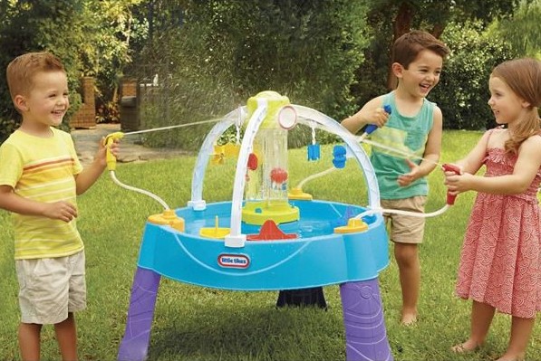 grup de trei copii jucandu-se in aer liber la o masuta de joaca dotata cu diverse dispozitive necesare jocului cu apa