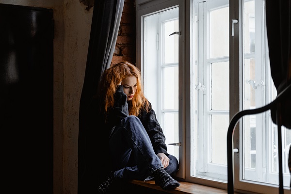 femeie tanara cu bluza neagra si blugi trista, stand ghemuita pe pervazul unei ferestre