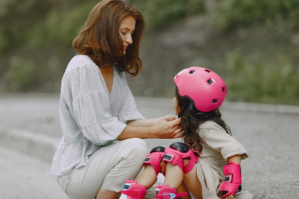 mama care sta pe vine pentru a-i fixa casca de protectie fetitei sale care merge cu rolele