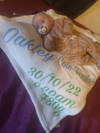 bebelus cu suzeta stand pe o paturica facuta special pentru el, cu numele sau complet si cu greutatea avuta la nastere