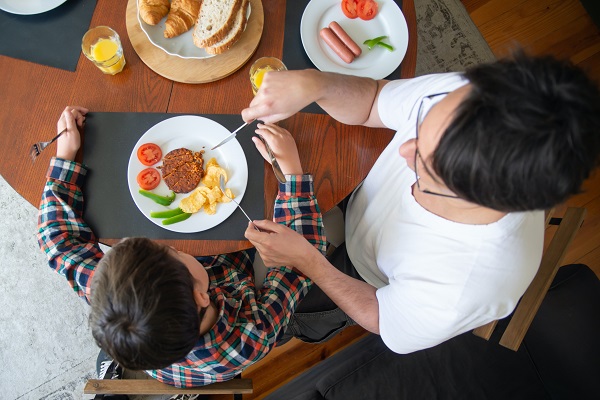 adult care sta la masa alaturi de un copil si il ajuta sa taie alimentele din farfurie 