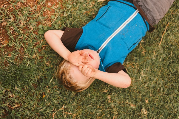 baietel cu tricou albastru care se freaca la ochi in timp ce sta intins pe iarba