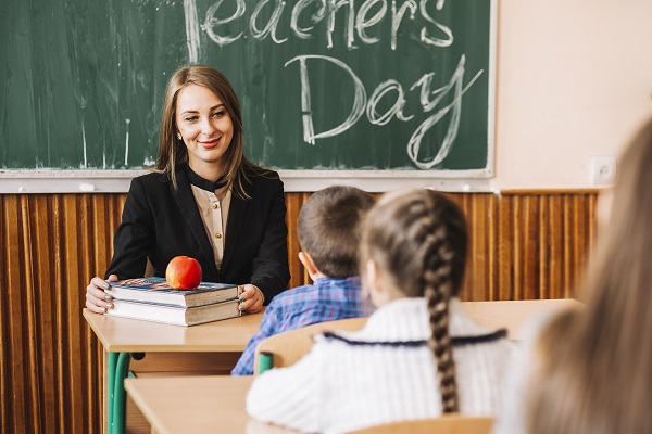 profesoara sau invatatoare zambitoare, stand la catedra si sarbatorind alaturi de elevii ei Ziua Profesorilor