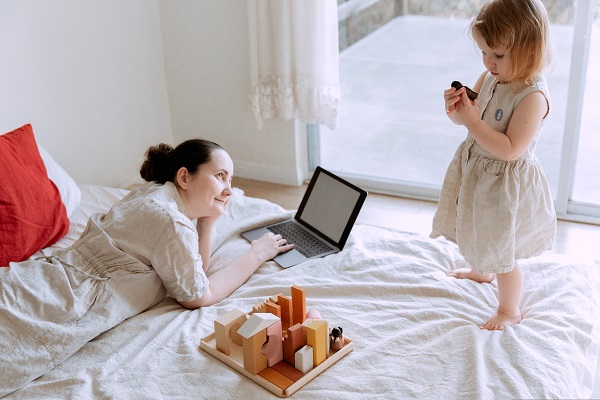 mama care lucreaza la laptop din pat, in timp ce isi priveste fetita care se joaca langa ea