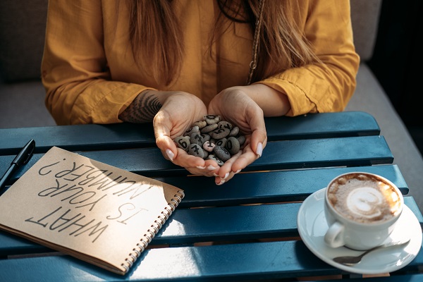 femeie care sta la masa pentru a servi a cafea si care tine in mana pietre cu mai multe cifre, intrebandu-se care este cifra destinului ei