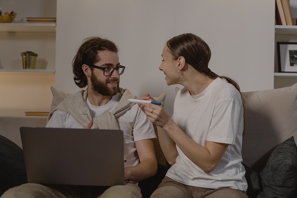 femeie entuziasmata care îi arata rezultatul pozitiv al testului de sarcina partenerului ei, care lucreaza pe laptop