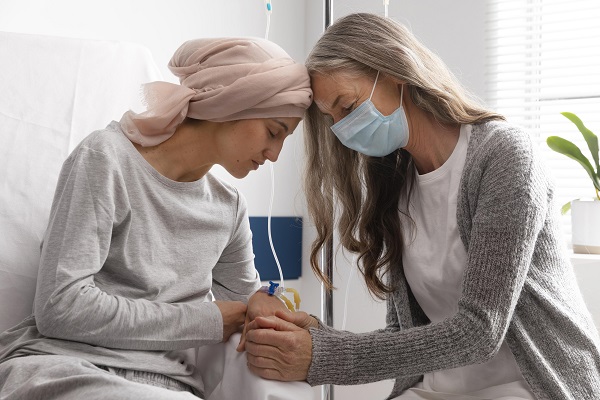 femeie matura cu masca de protectie incurajand o tanara pacienta bolnava de cancer, aflata pe patul de spital