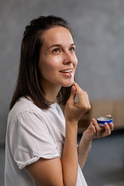 femeie tanara care aplica pe buze, cu un betisor, balsam de buze dintr-o cutiuta rotunda
