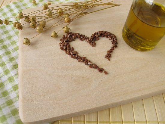 recipient de sticla cu ulei de in si seminte de in asezate in forma de inima, pe un tocator de lemn