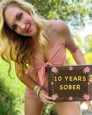 femeie tanara, blonda, fericita, tinand in mana o tablita pe care scrie „10 ani de cand sunt treaza”
