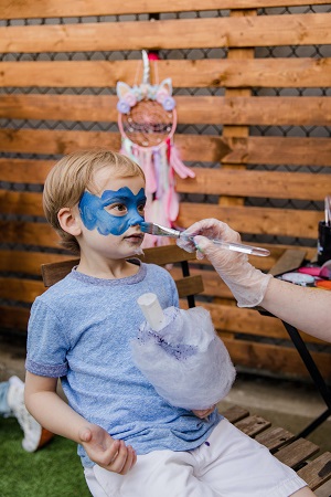 persoana care îi face o pictura pe fata, cu albastru, unui baietel