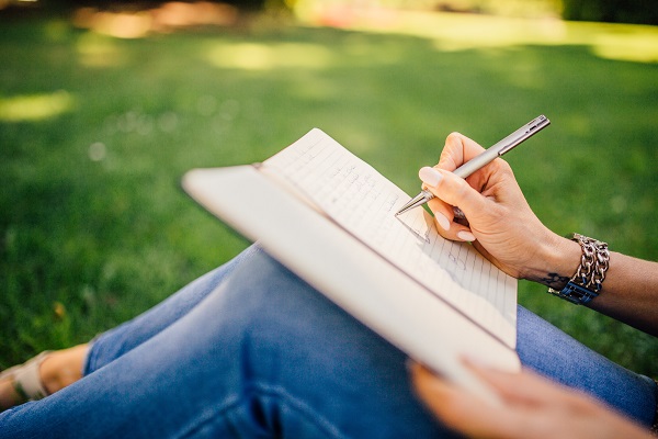 femeie in blugi, care sta pe iarba si tine pe genunchi un jurnal in care noteaza ceva
