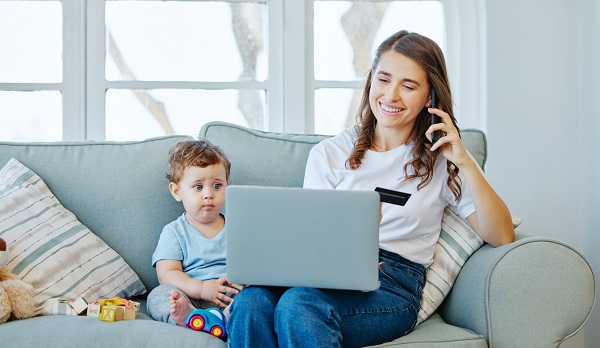 mama vesela care vorbeste la telefon, care tine in mana un card și se uita pe laptop avandu-l langa ea pe baietelul ei