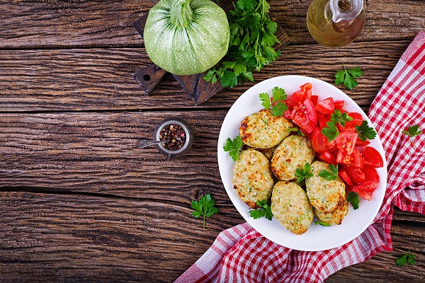 chiftele din dovlecei pe o farfurie alba, alaturi de  felii de rosii decorate cu frunze de patrunjel