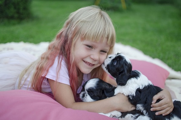 fetita cu parul lung si blond, cu nuante de roz, stand pe burta pe iarba in timp ce doi catelusi o ling pe față