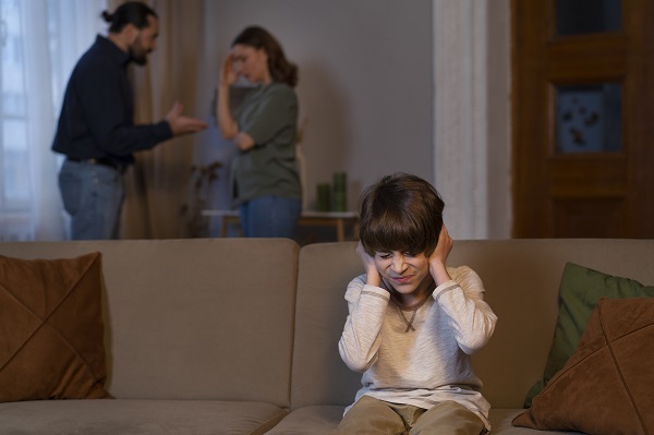 baietel care sta pe canapea si isi astupa urechile pentru a nu mai auzi cearta dintre parintii sai