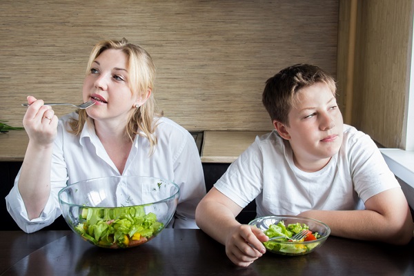 mama care mananca dintr-o salata, in timp ce fiul ei sta langa ea ingandurat