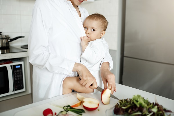 mama care taie mere in bucatarie, in timp ce isi tine bebelusul la san ca sa il alapteze