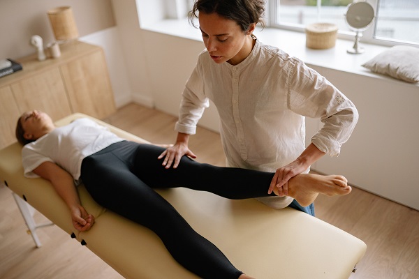 femeie tanara intinsa pe pat, in timp ce kinetoterapeuta lucreaza cu unul dintre picioarele ei