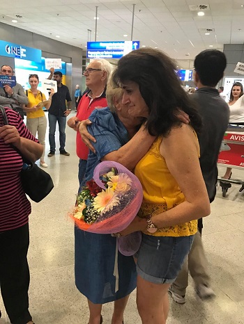 femeie in pantaloni scurti de blugi, care imbratiseaza o femeie in varsta, ce tine un buchet de flori, in aeroport