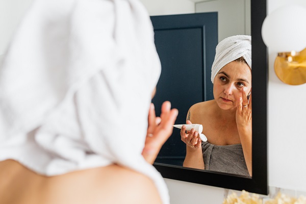 femeie infasurata cu un prosop gri, stand in fata oglinzii din baie si aplicand pe fata o crema