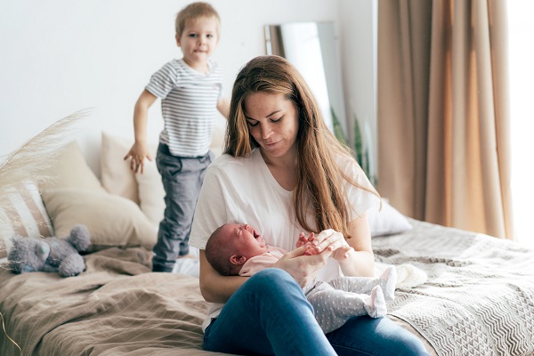 mama care isi tine in brate bebelusul care plange, in timp ce baietelul ei mai mare sare pe pat