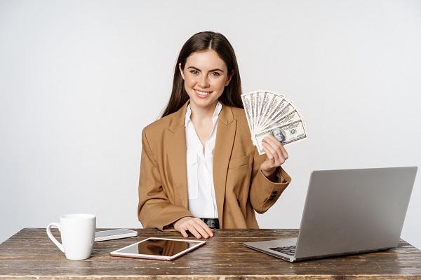 femeie in tinuta office fericita, in timp ce tine in mâna bancnote si se afla in fata laptopului