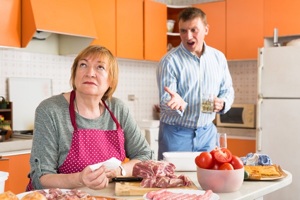 barbat tanar, care tine in mâna un pahar cu bautura si care se cearta cu o femeie matura, care gateste un fel de mancare cu carne