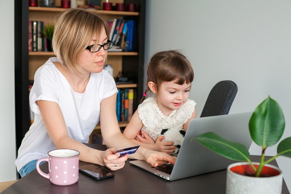 mama in fata unui laptop, incercand sa faca o plata online cu cardul in timp ce fetita ei sta langa ea
