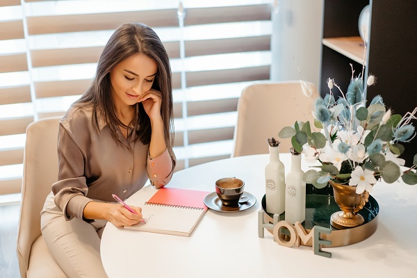 femeie purtand bluza si pantaloni in nuante de crema, stand relaxata la biroul de acasa si notandu-si ceva intr-un carnet