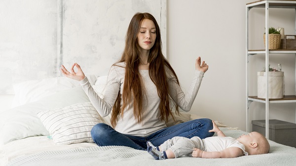 mama care sta pe pat si mediteaza cu ochii inchisi, in pozitie de yoga, in timp ce bebelusul ei se afla langa ea