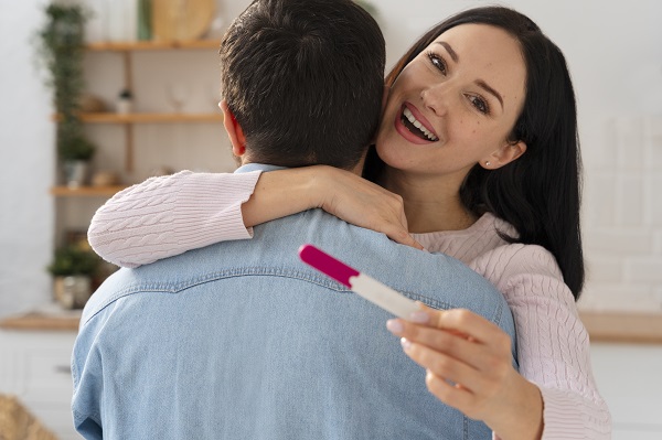 femeie fericita, care tine in mana un test de sarcina si isi imbratiseaza partenerul