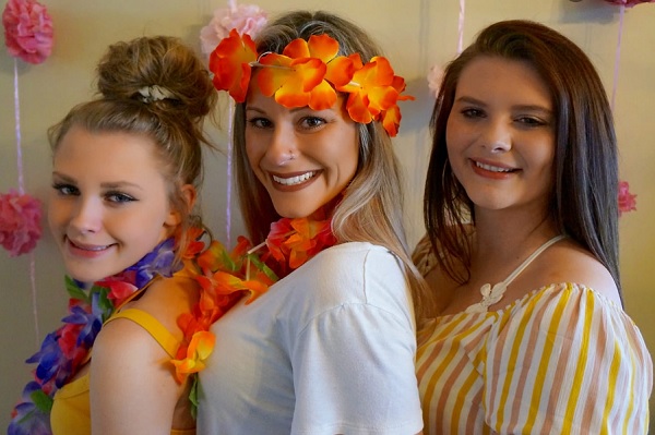 femeie matura si doua adolescente fericite, mama purtand pe cap o coronita din flori colorate, iar una dintre fiicele ei un colier din astfel de flori