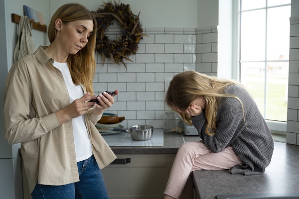 mama care se uita pe telefon si fetita trista, in bucatarie