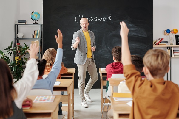 profesor care sta la tabla si le preda elevilor sai, care stau cu mainile ridicate sa raspunda