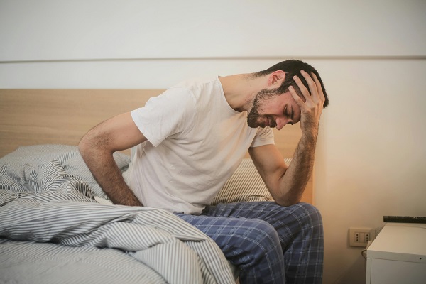 barbat in pijama care sta pe marginea patului intrucat nu se simte bine din pricina unei dureri de cap