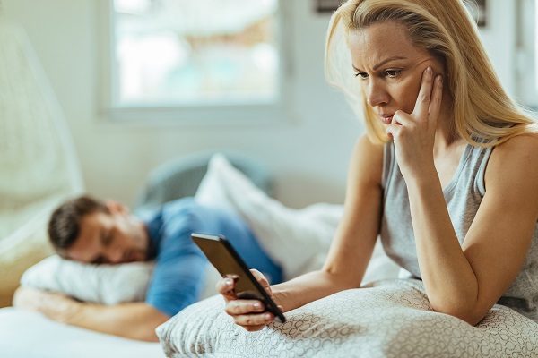 femeie trista care se uita pe telefon in timp ce partenerul ei doarme