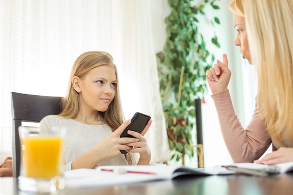 adolescenta care sta la masa cu telefonul si mama care incearca sa o disciplineze