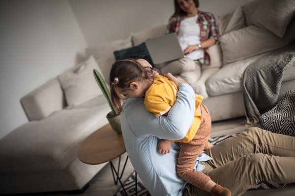 tata care isi imbratiseaza fetita, iar pe fundalul imaginii se vede mama care sta pe canapea cu laptopul