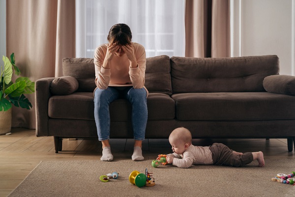 mama disperata, stand pe canapea cu fata intre maini, in timp ce bebelusul ei se joaca pe covor