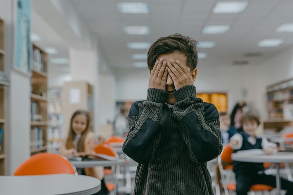 baietel care isi acopera fata cu mainile si sta cu spatele la colegii de clasa