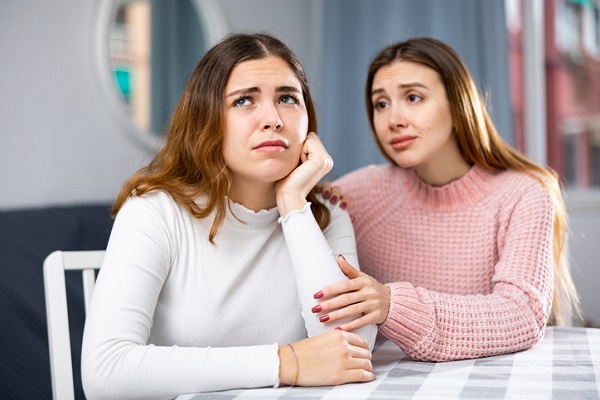 femeie tanara care incearca sa o consoleze pe alta femeie tanara, ambele stand la masa