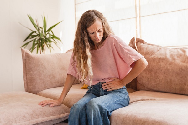 femeie cu parul lung, care sta pe canapea si isi tine mana pe o parte a abdomenului inferior din cauza disconfortului sau durerilor