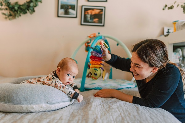 bebelus care sta pe burtica sprijinindu-se de o perna si mama care se joaca cu el aratandu-i o jucarie colorata