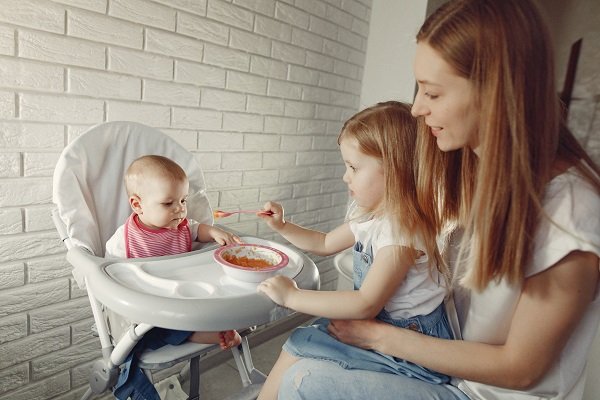 surioara mai mare care incearca sa-si hraneasca fratiorul bebelus cu hrana pasata din castronel